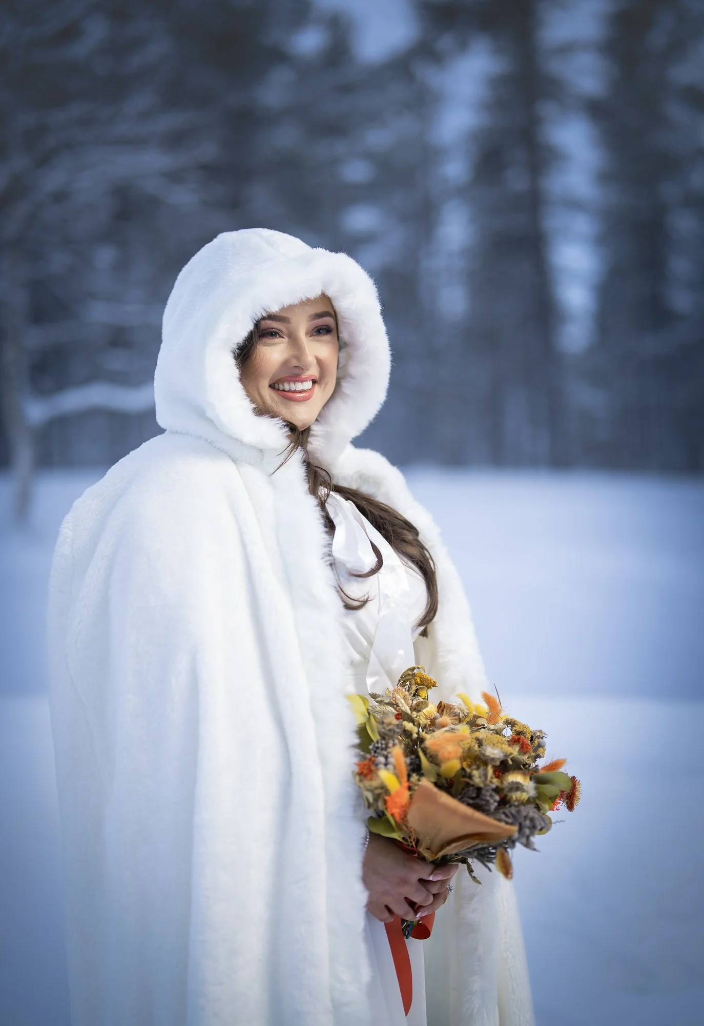A woman dressed in a white winter cloak with a hood standing outdoors in a snowy forest, smiling and holding a bouquet of flowers.