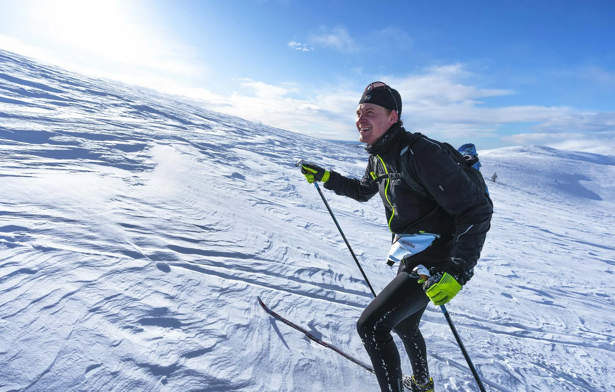 A man skiing on a snowy mountain slope under a partly cloudy sky, smiling and wearing black and green skiing gear.
