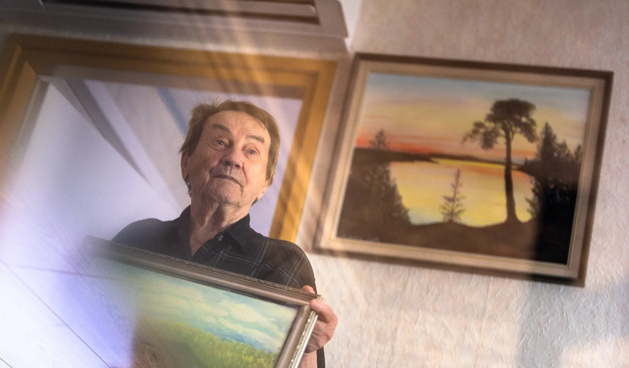 An elderly man with light brown hair, wearing a dark plaid shirt, holds a landscape painting. The painting depicts a green field with trees in the distance and a blue sky. The photo is taken from below, with the man reflected in a mirror, and there a