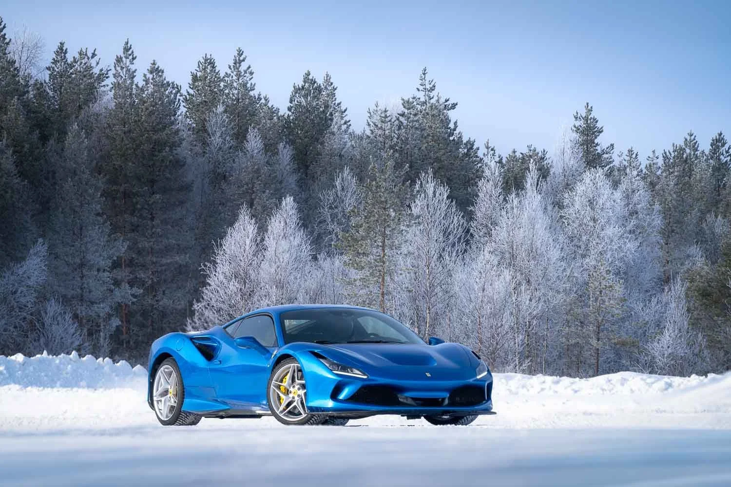 A blue sports car parked on snow-covered ground with snow-covered trees and a clear blue sky in the background.