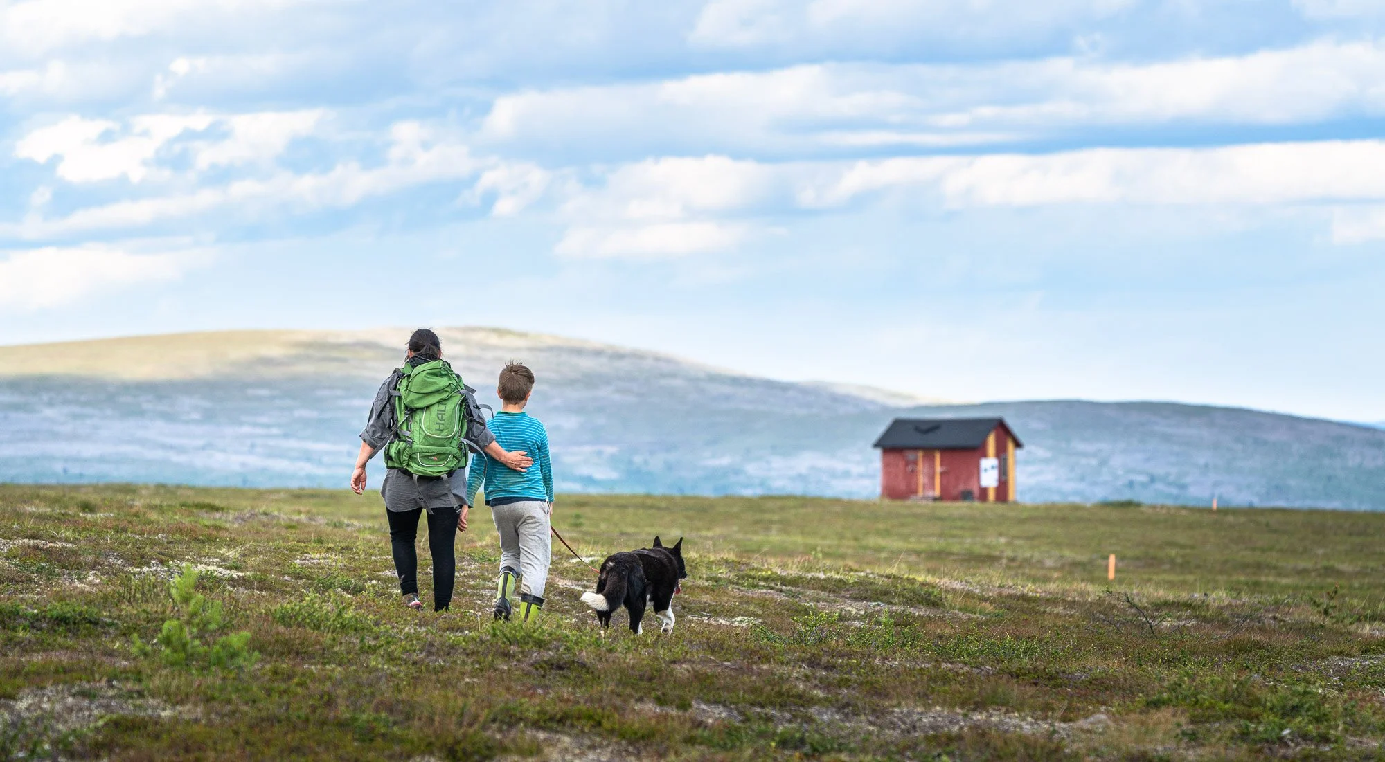 A woman and boy walking through a green field with a dog, with a small red house in the distance under a partly cloudy sky.