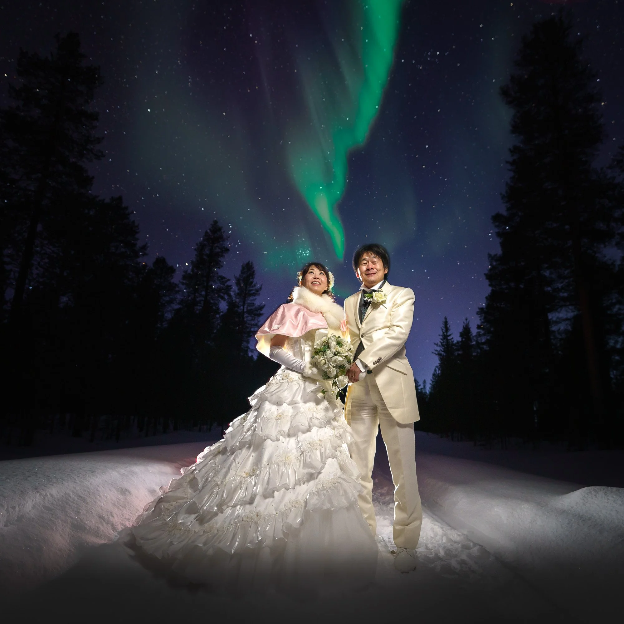 A newlywed couple in wedding photoshoot in Saariselkä standing on snow under a starry sky with the Northern Lights overhead.