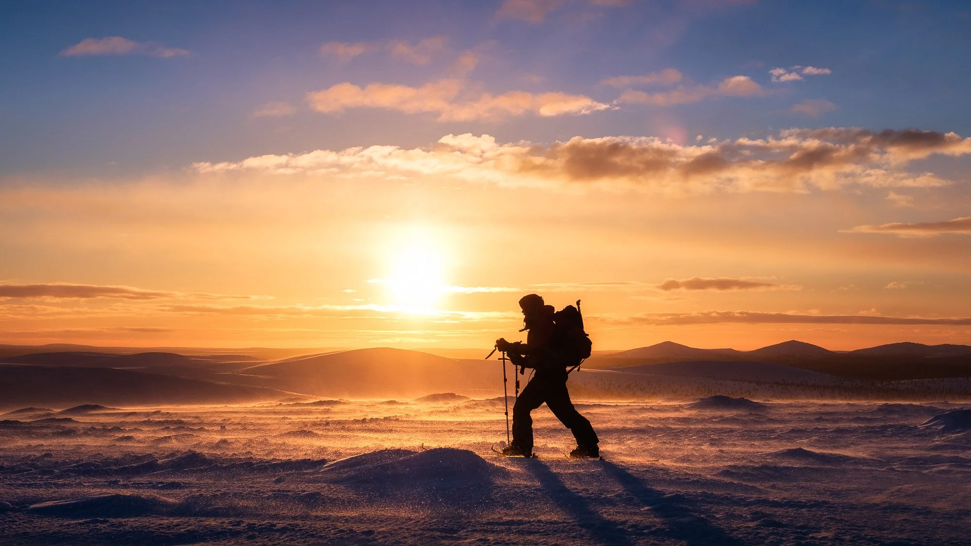 A person snowboarding on a snowy landscape during sunset in Kiilopää, Saariselkä, with a backpack and trekking poles, under a colorful sky with clouds.