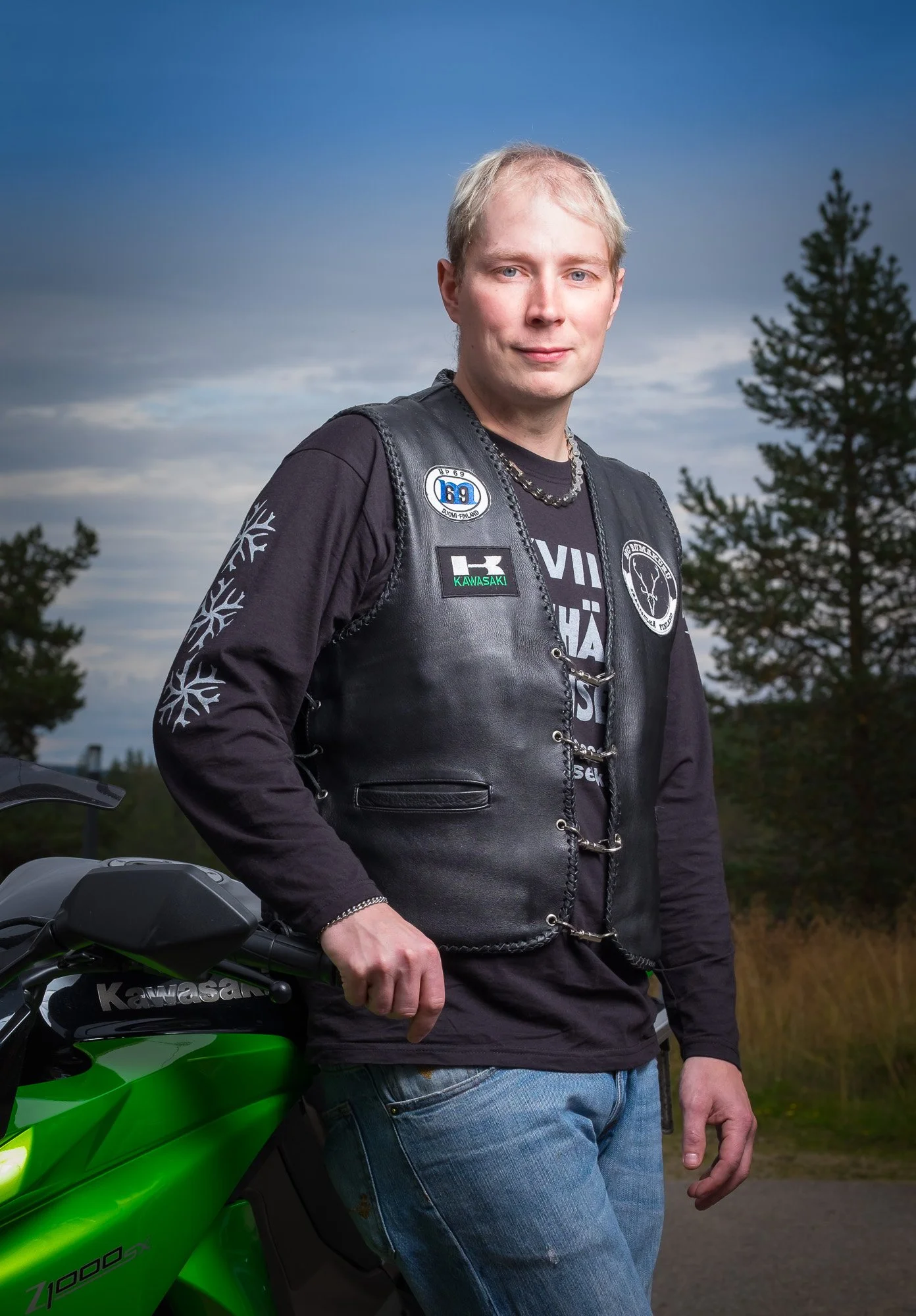A man with blond hair standing outdoors in front of a mountain landscape and trees, leaning on a green Kawasaki motorcycle, wearing a black leather vest and long-sleeve shirt.