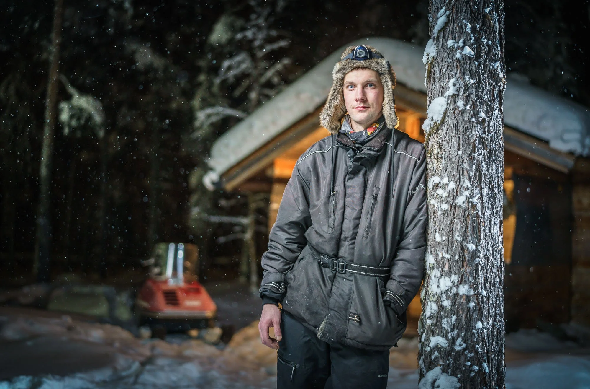 A man in winter gear leaning against a snow-covered tree at night, standing outside a wooden cabin with warm lights in a snowy forest.