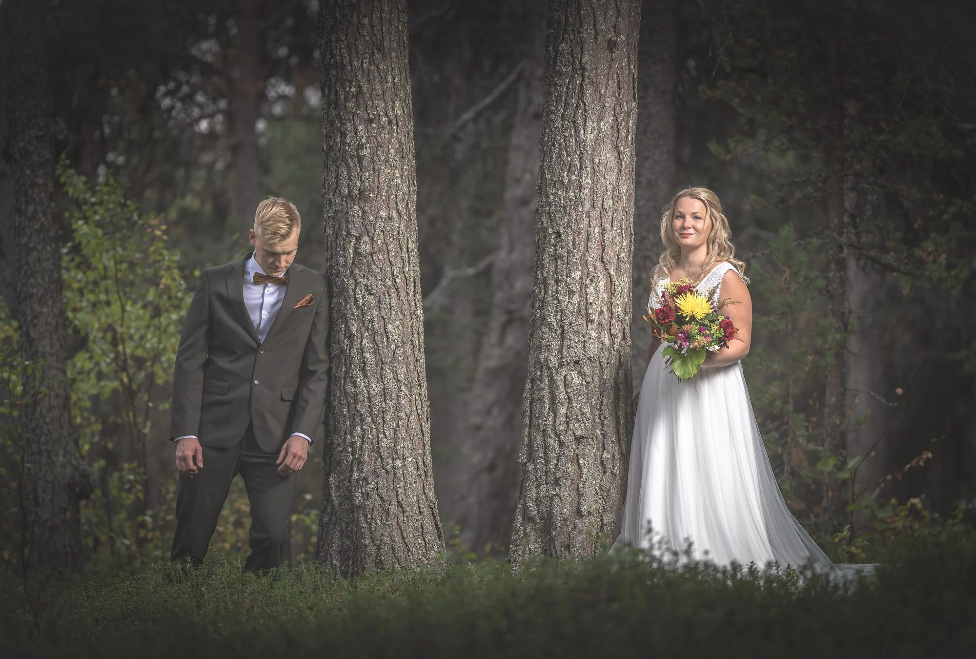 A bride in a white wedding dress holding a bouquet of yellow, red, and purple flowers, standing behind a tree, looking at the camera. A groom in a black suit with a brown bow tie stands behind another tree, looking down, in a forest setting.