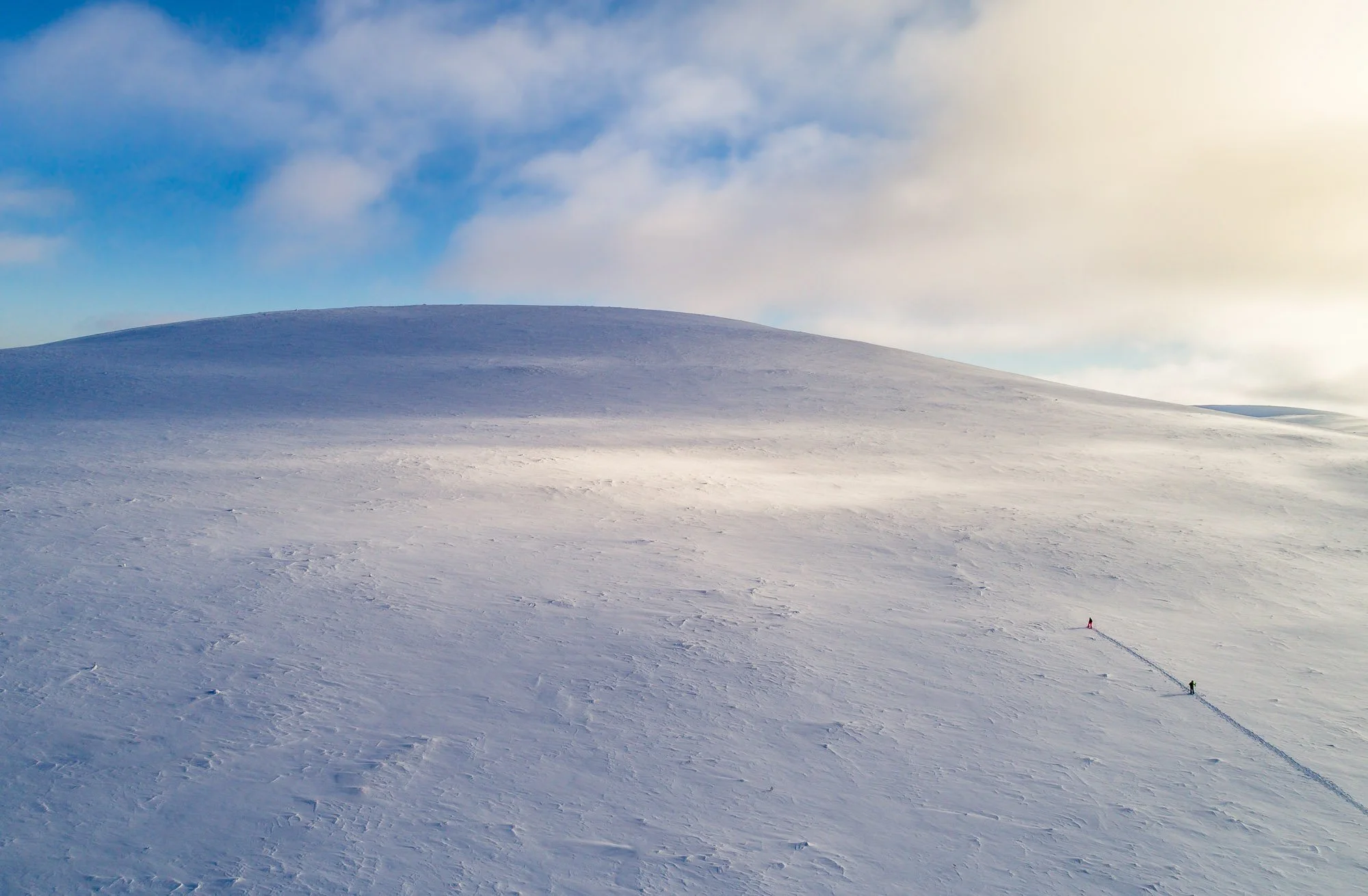 Snow-covered mountain with two small figures hiking and leaving a trail behind, under a cloudy sky with some sunlight breaking through.