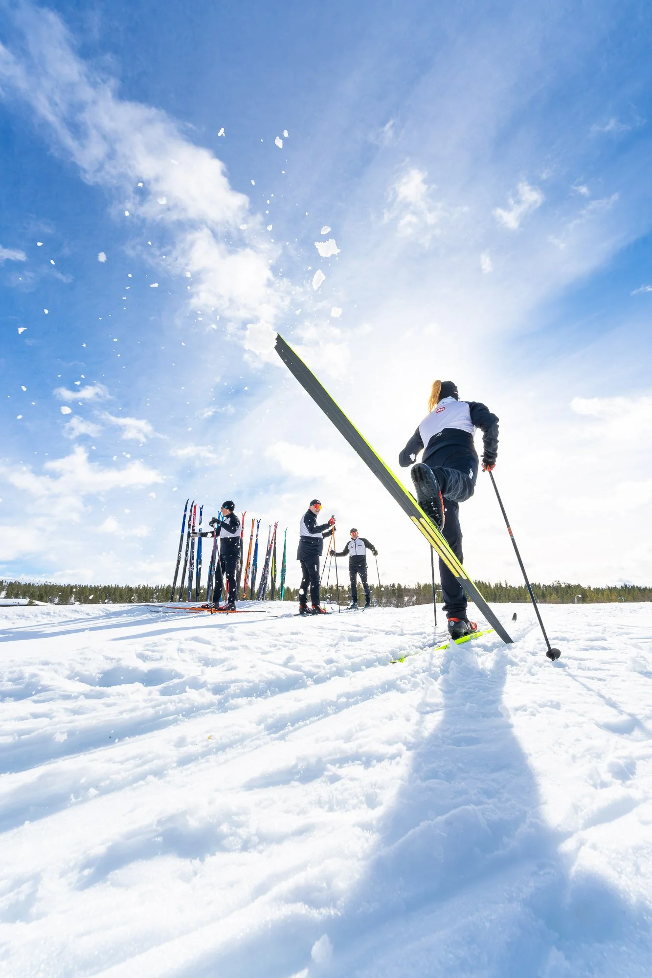 People cross-country skiing on a snowy field under a blue sky with clouds, with some skis lined up in the background.