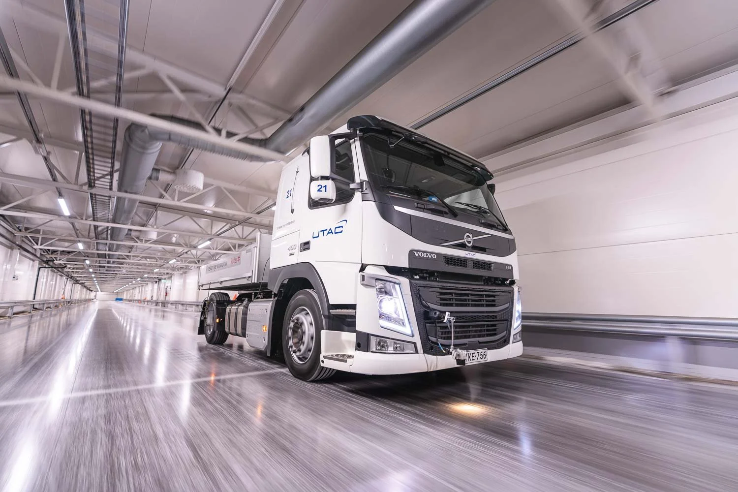 A white Volvo truck with the logo 'UTAC' on the side, driving through a tunnel with metallic pipes on the ceiling and bright overhead lighting.