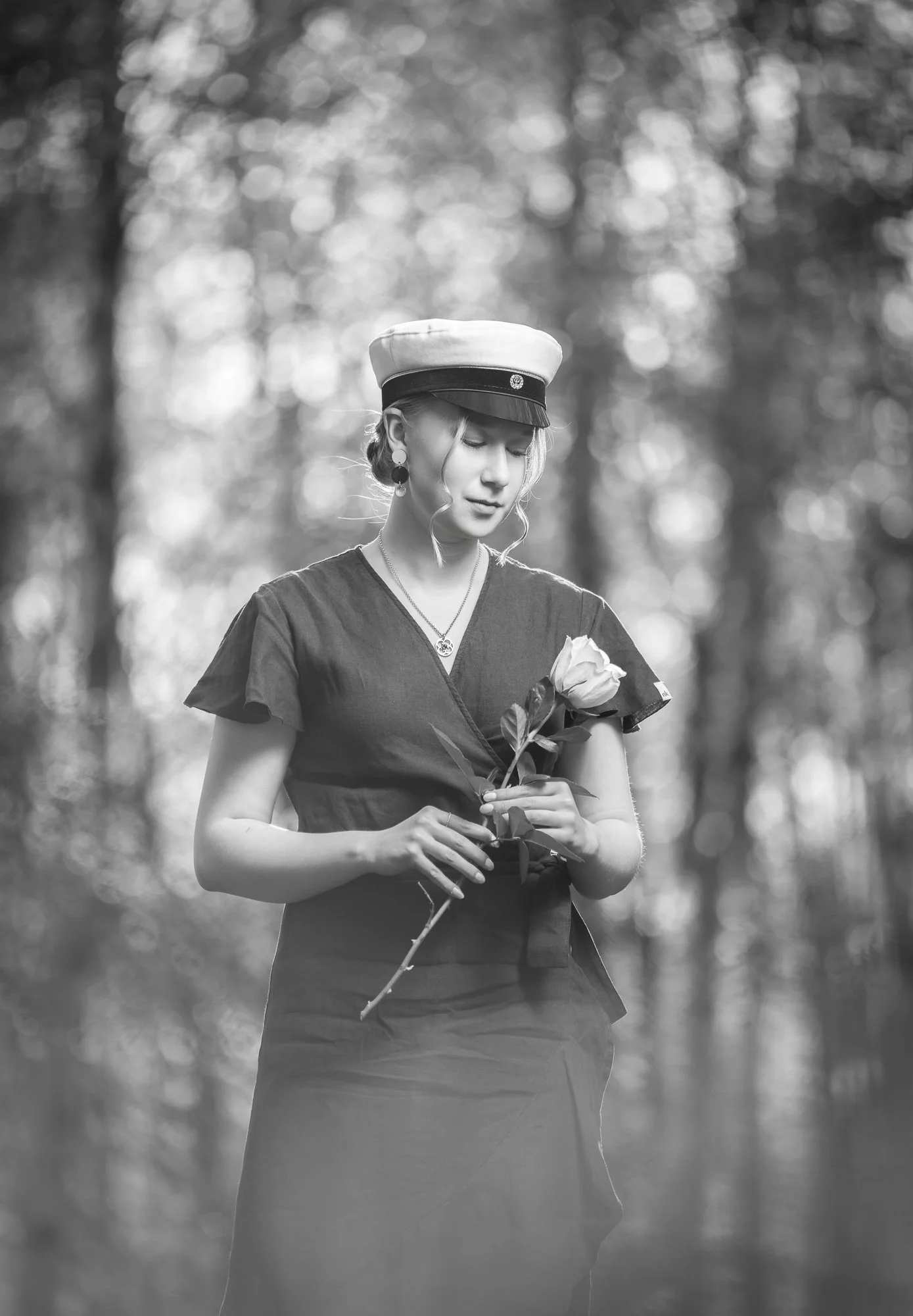 A woman standing outdoors in a forested area, holding a rose, wearing a military-style hat and dark clothing, with her eyes closed and a serene expression.