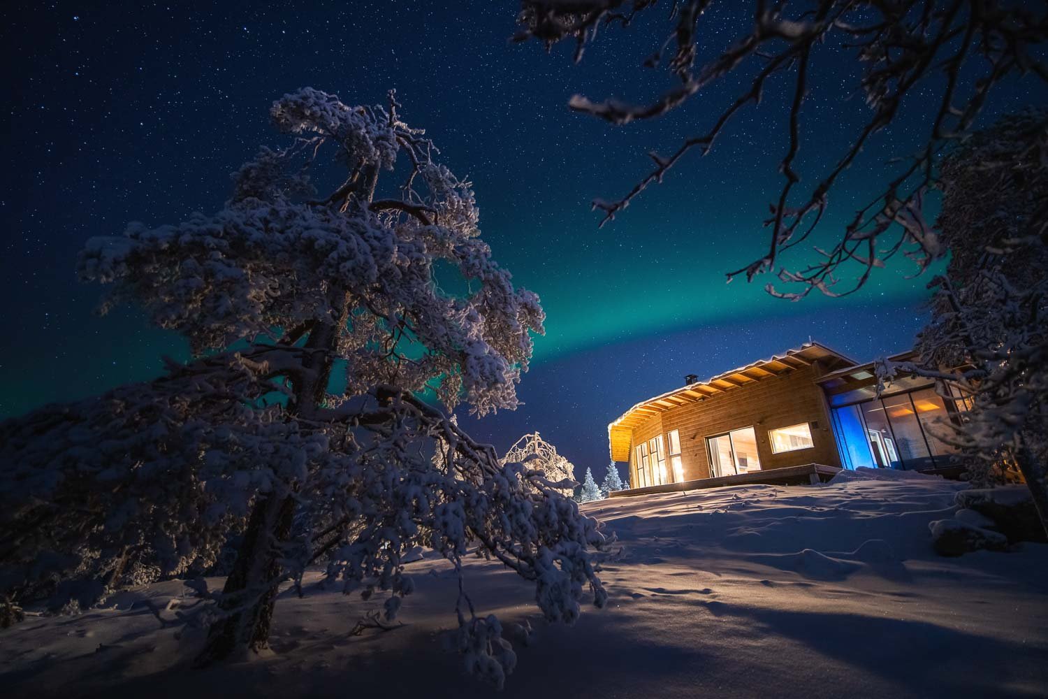 A snowy landscape at night with a modern house illuminated from within, snow-covered trees, and the Northern Lights in the sky.
