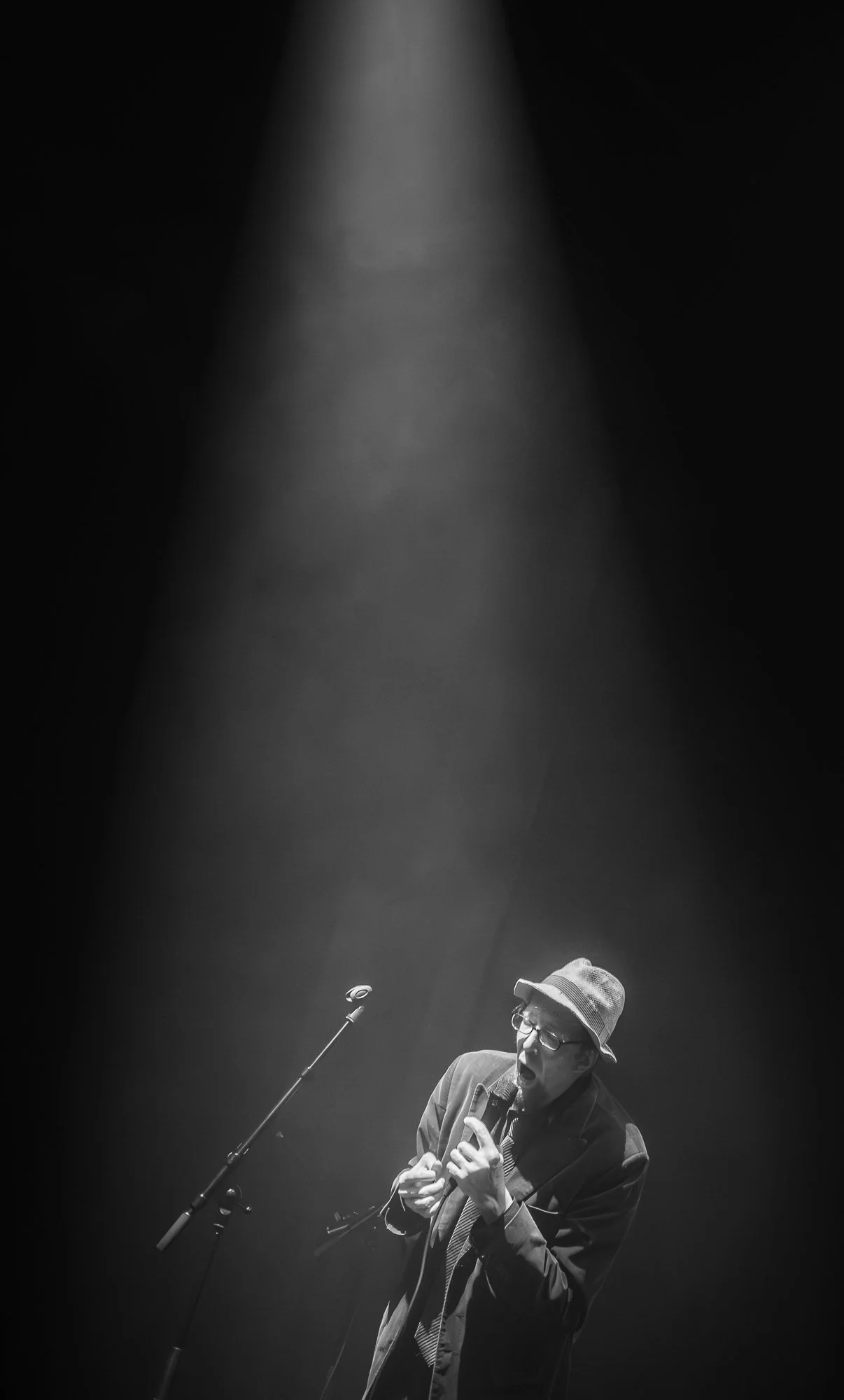 A black and white photo of a man wearing glasses, a hat, and a suit, singing into a microphone on stage, illuminated by a spotlight.