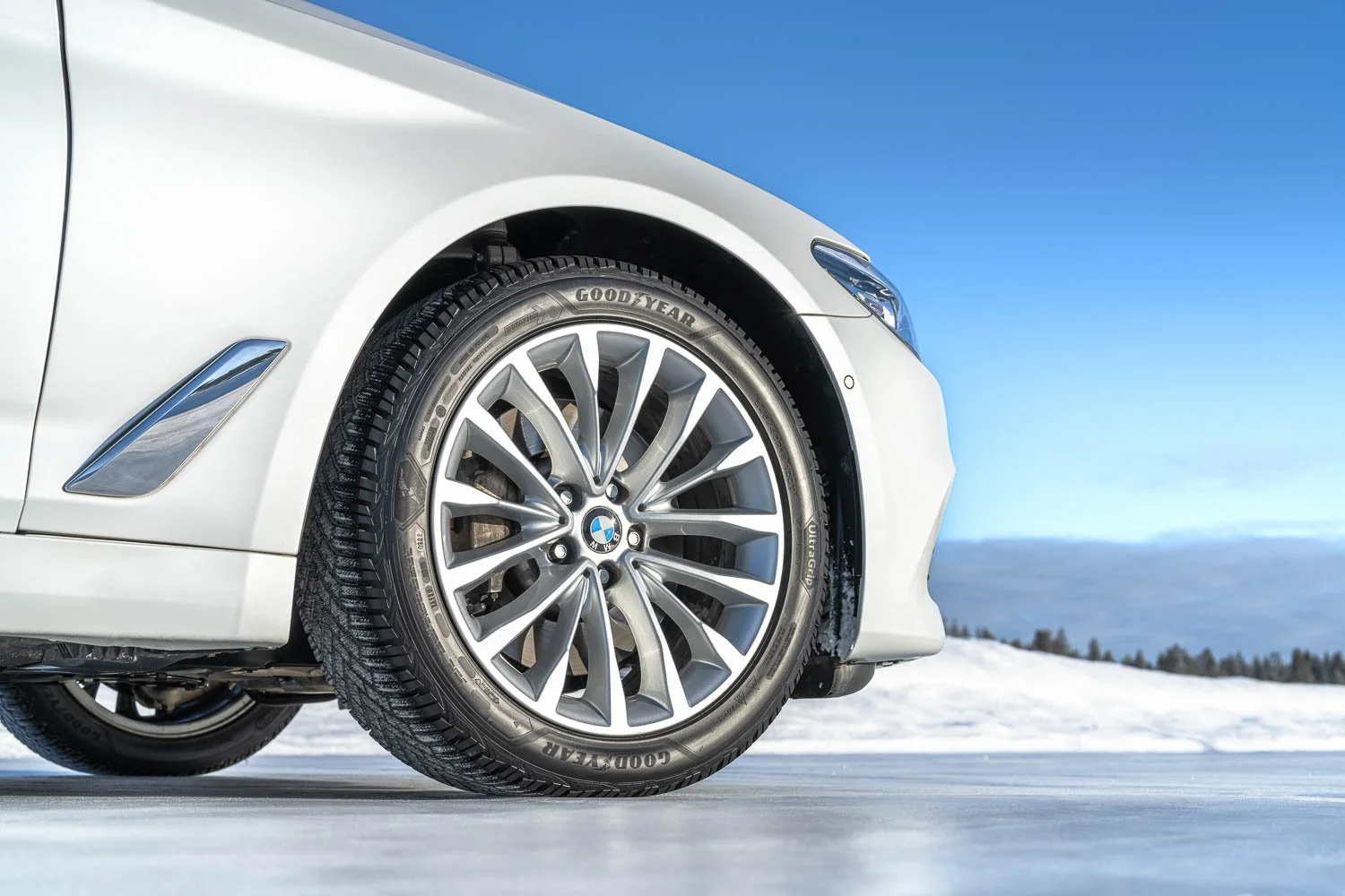 Close-up of the front left wheel of a white BMW car parked on snow-covered ground against a blue sky and distant trees.
