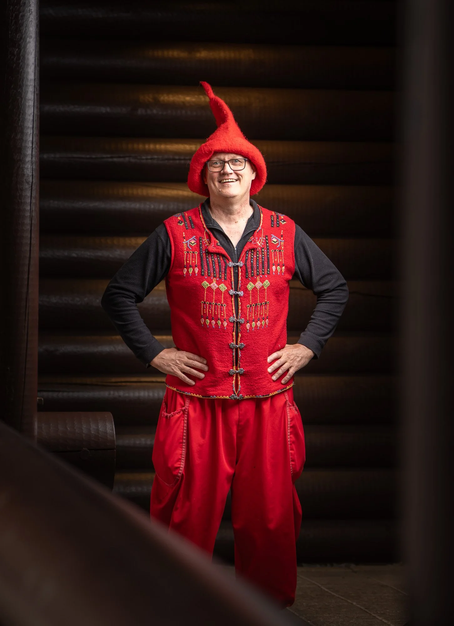 A man dressed in a red Viking helmet, red embroidered vest, and red baggy pants stands with hands on hips, smiling in front of a dark wooden staircase.