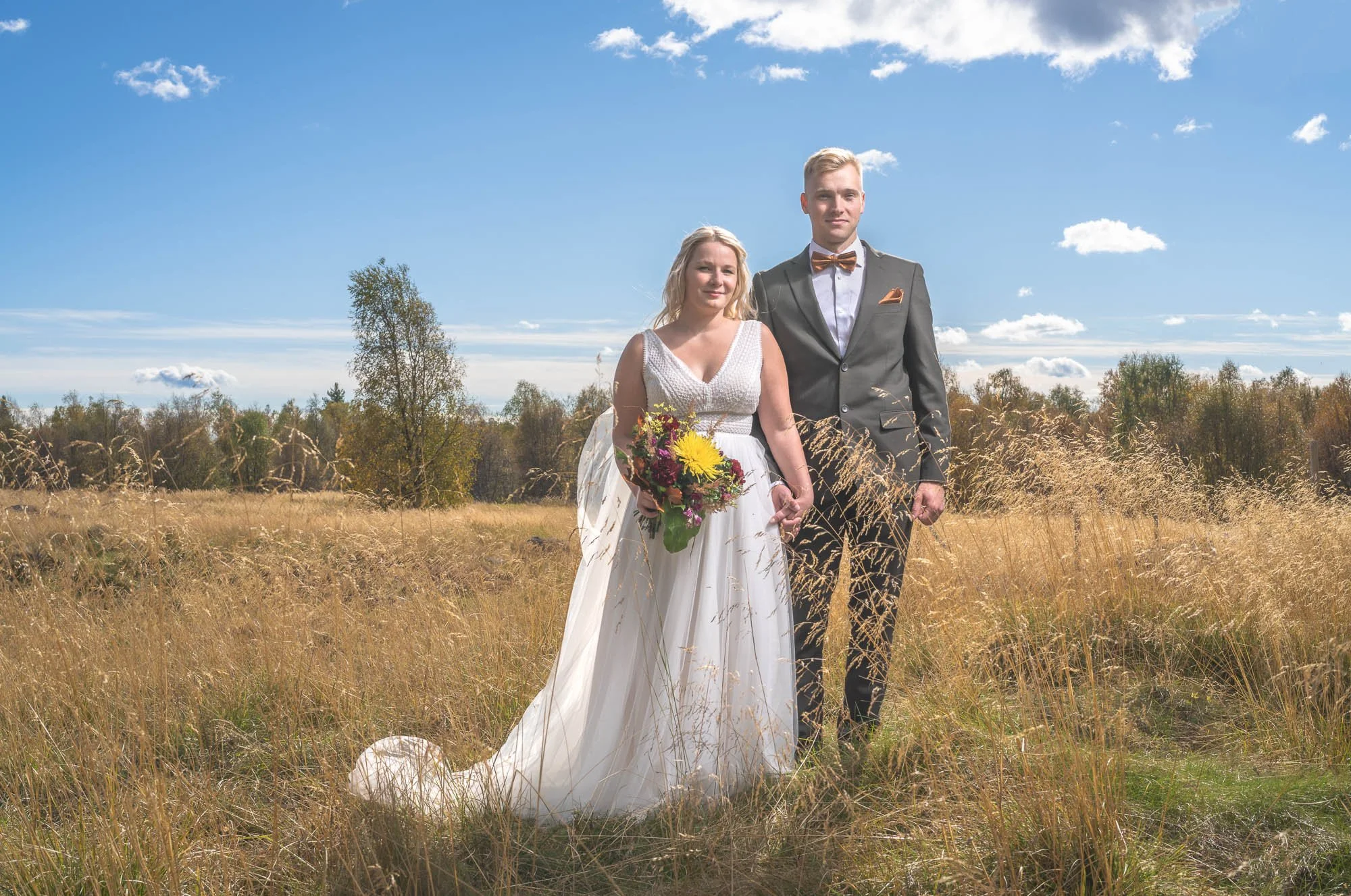 A bride and groom standing in a grassy field on a sunny day, with the bride holding a bouquet of flowers.
