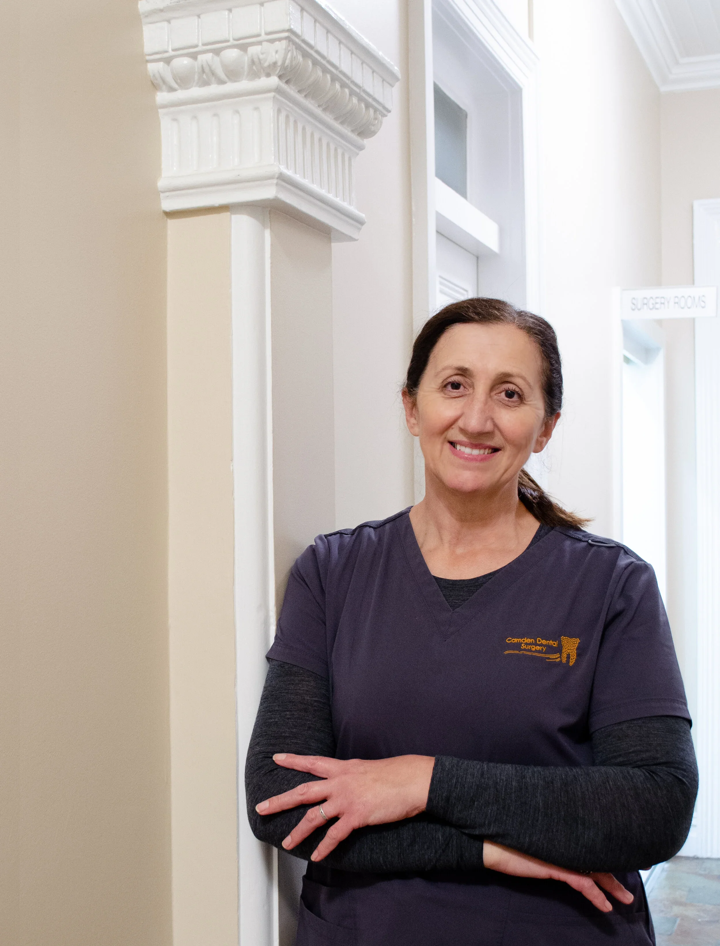 A woman in a dark purple dental scrubs top, smiling, standing against a wall in a healthcare facility. A sign in the background reads "Surgery Rooms."