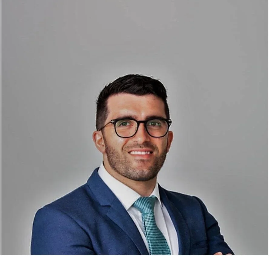 A professional portrait of a man with short dark hair, glasses, and a beard, wearing a blue suit, white shirt, and a teal tie, standing against a plain grey background.