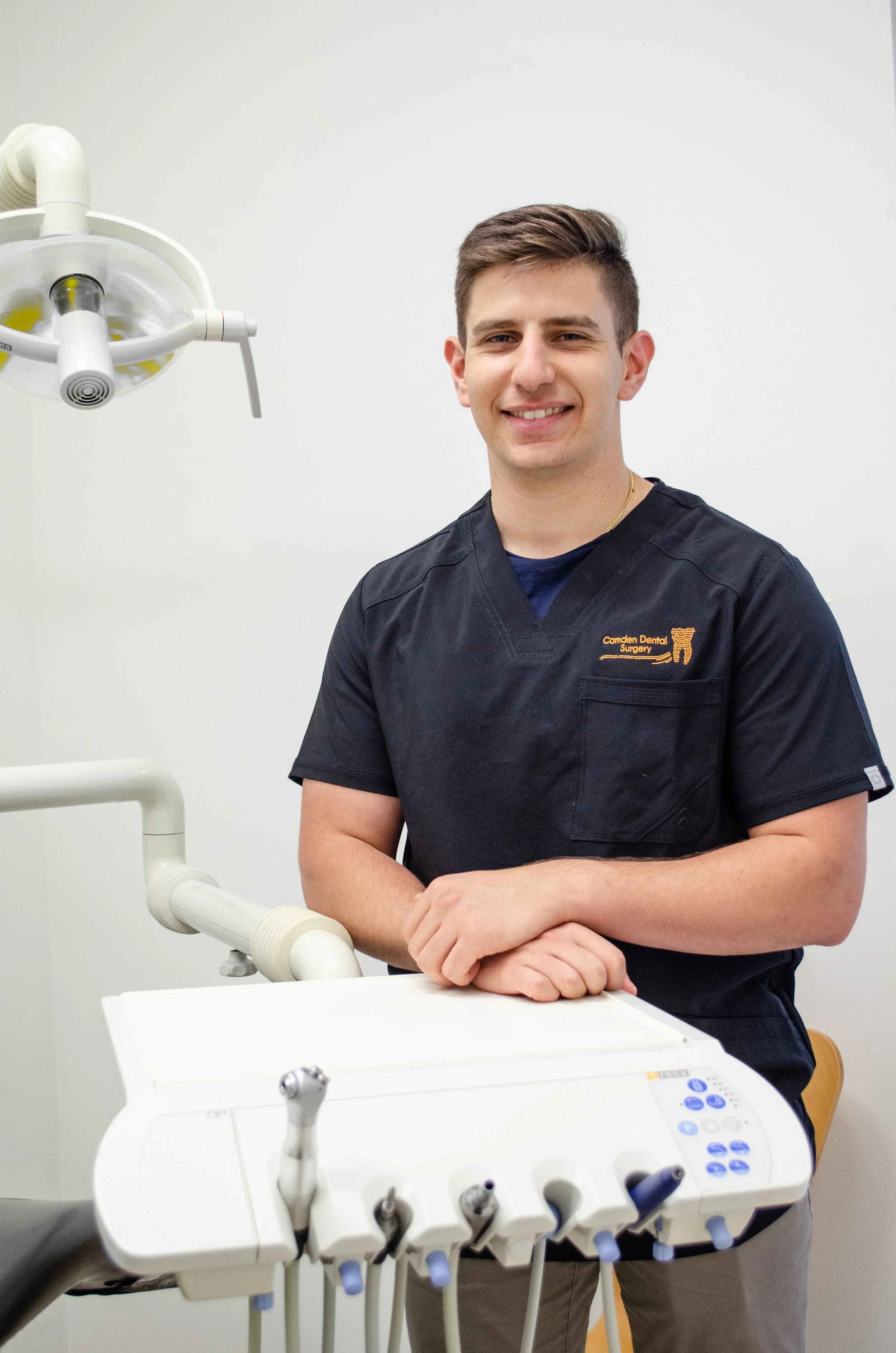 A young man in a black dental uniform standing next to dental equipment in a clinical setting, smiling at the camera.
