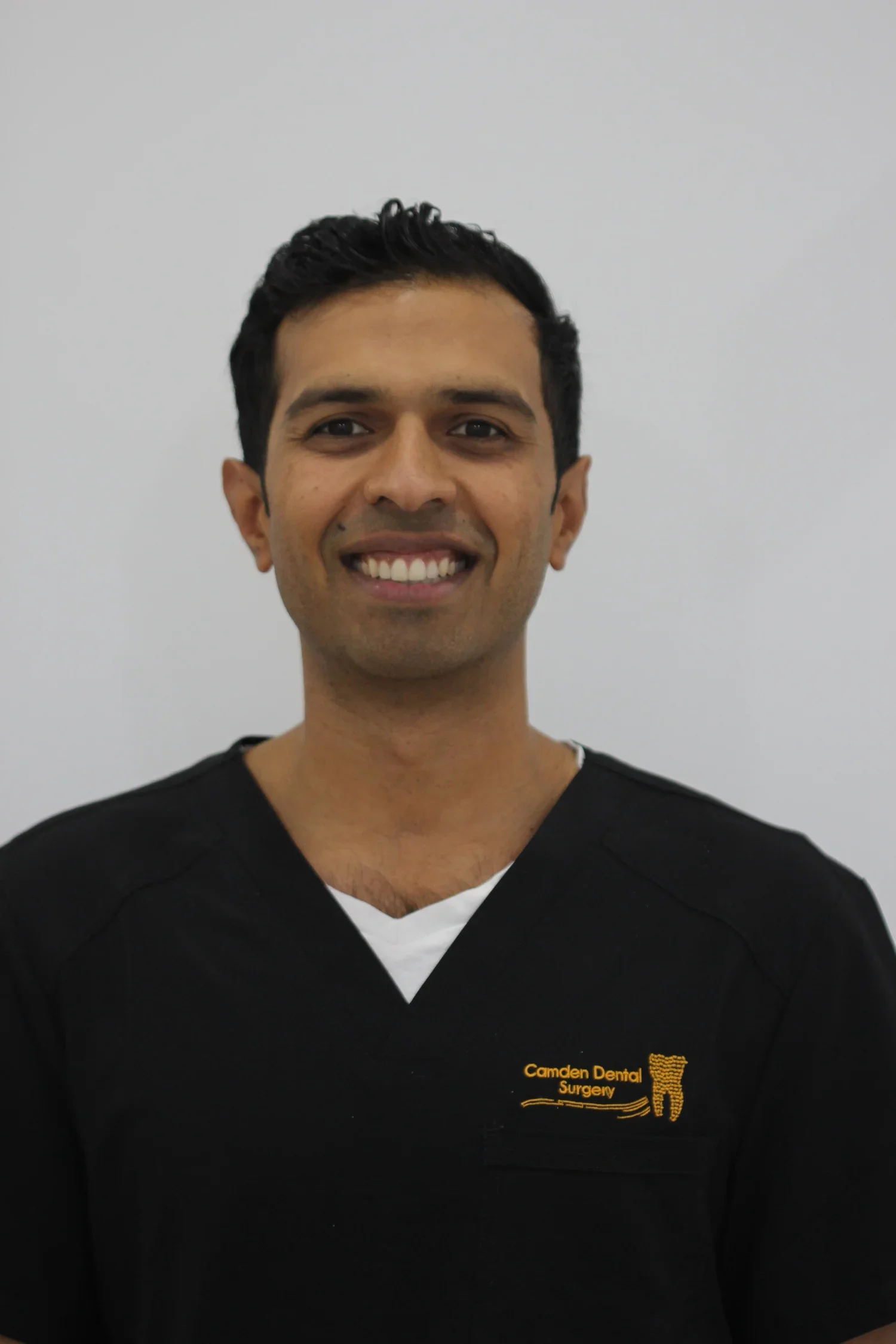 A smiling man wearing a black dental uniform with a logo that reads 'Camden Dental Surgery' and features a tooth graphic, standing against a plain light background.