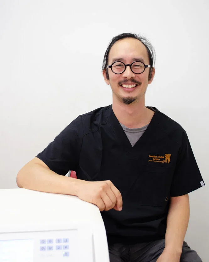 A man with glasses and facial hair wearing a black medical uniform, sitting at a desk with a white background.