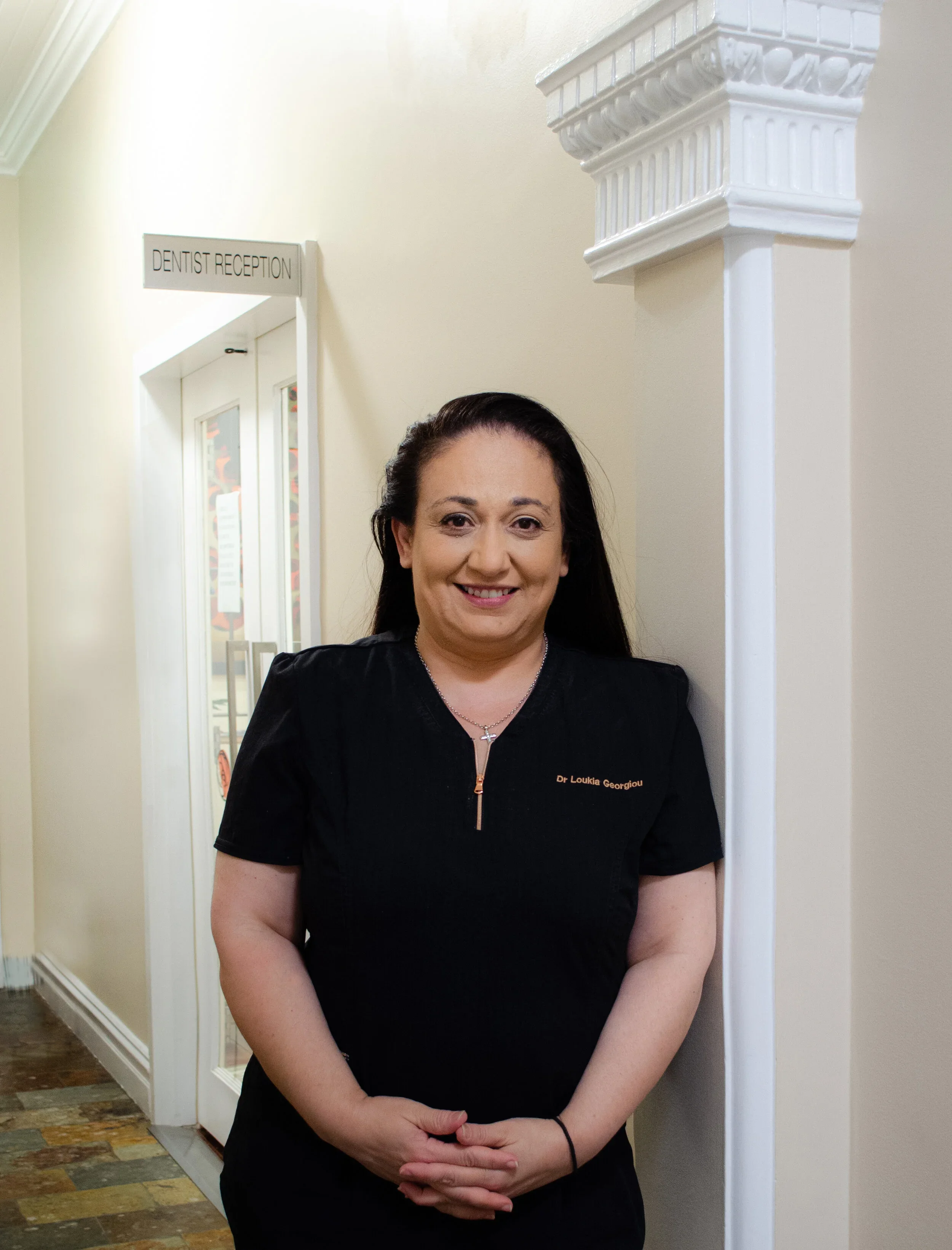 A woman in black scrubs with the name tag 'Dr. Loukia Georgiou' standing indoors next to a wall with a decorative column. Behind her is a door with a sign that reads 'DENTIST RECEPTION'.