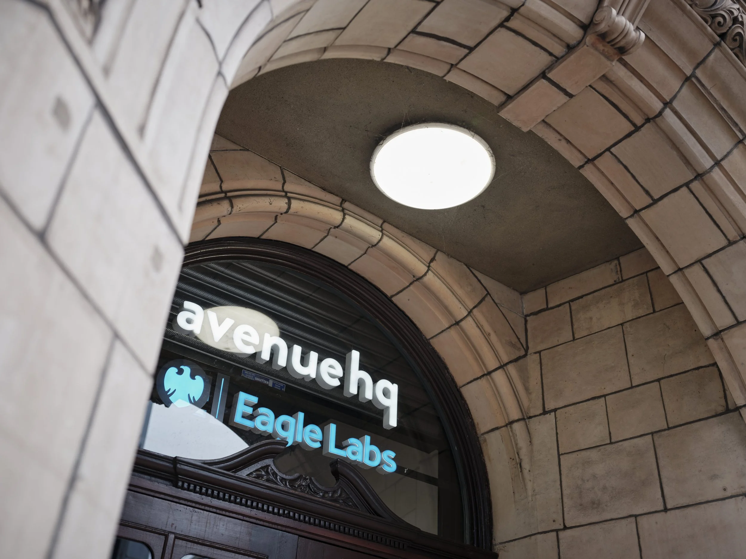 View of a building entrance with the signs 'avenueh' and 'Eagle Labs' above the door. The entrance is framed with stone bricks and a circular ceiling light is visible.