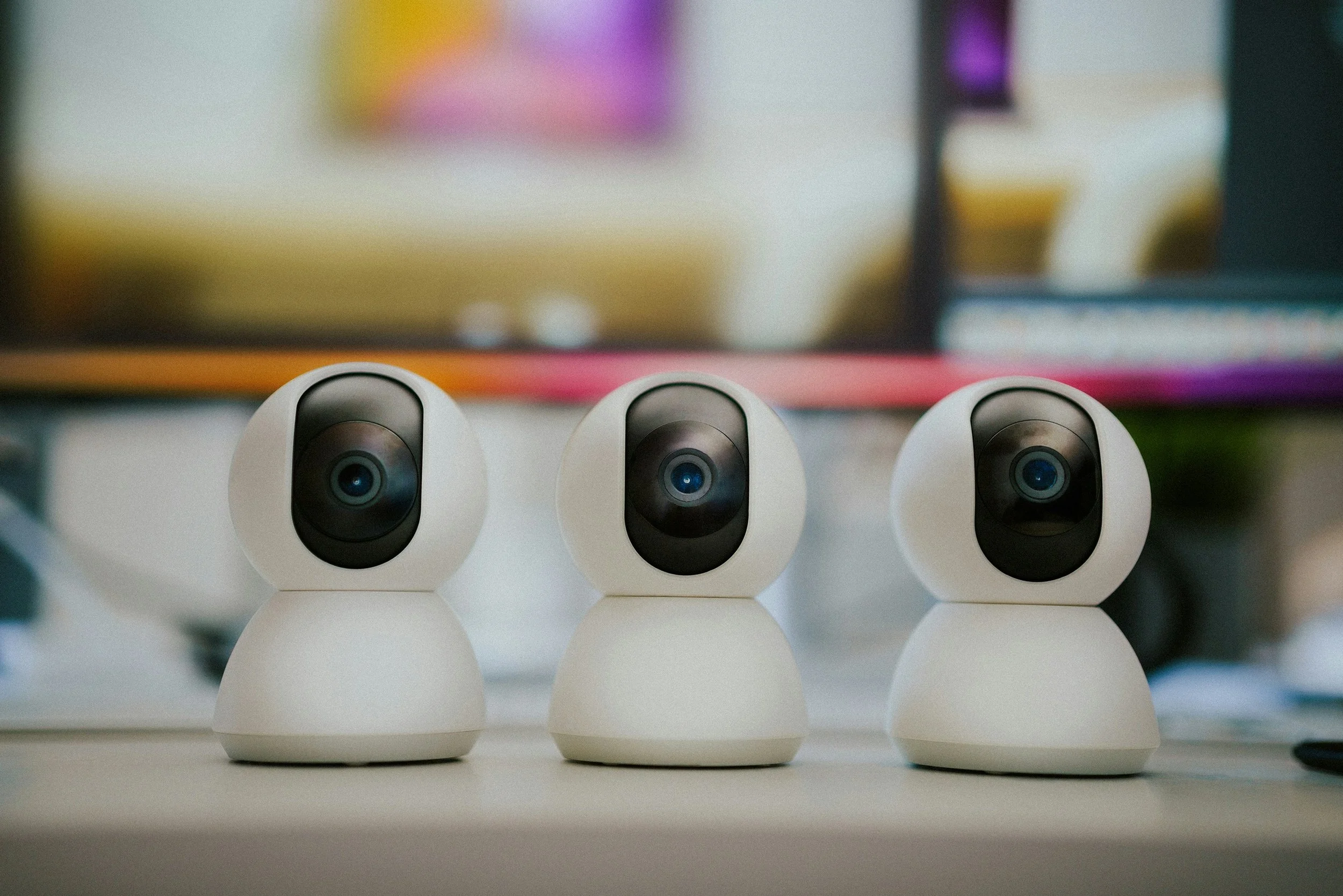 Three white, robot-like surveillance cameras with black lenses are placed on a white surface with a blurred background.