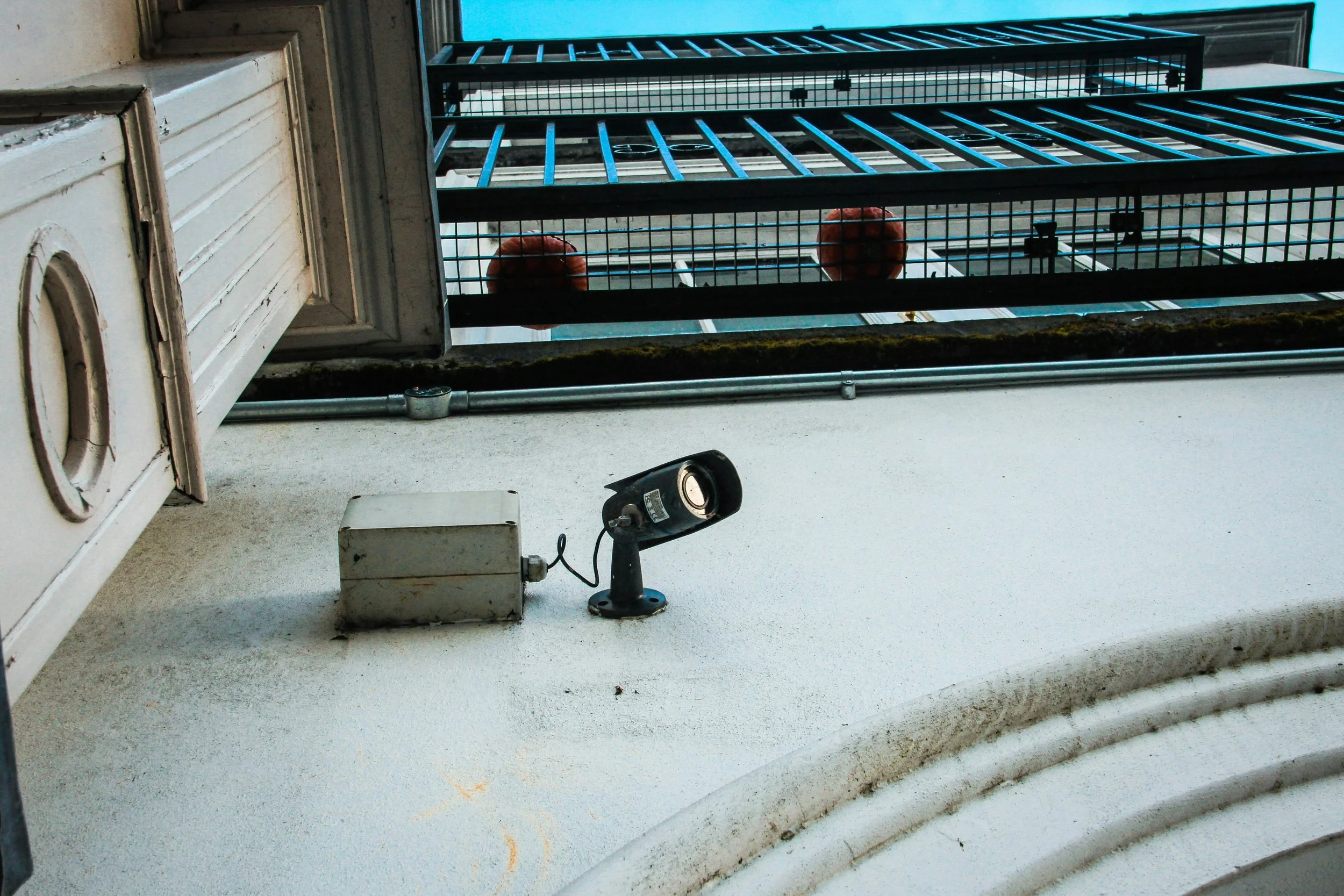 Looking up at a building's exterior wall shows a security camera, sign of rust and moss near the roofline, and a fire escape with two red fire extinguishers visible through a metal grid.