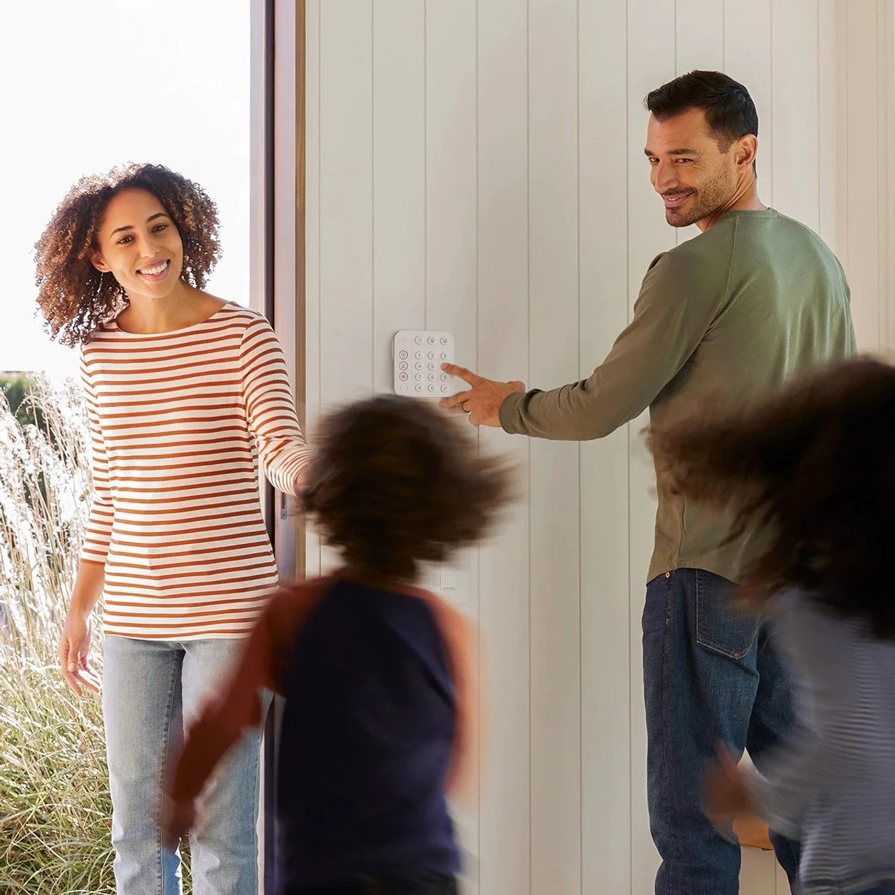 A man presses a doorbell button as a woman and children enter a house, smiling.