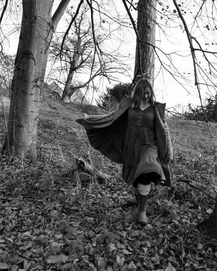 A woman walking through a wooded area on a slope, with leafless trees around her and fallen leaves on the ground, in black and white.
