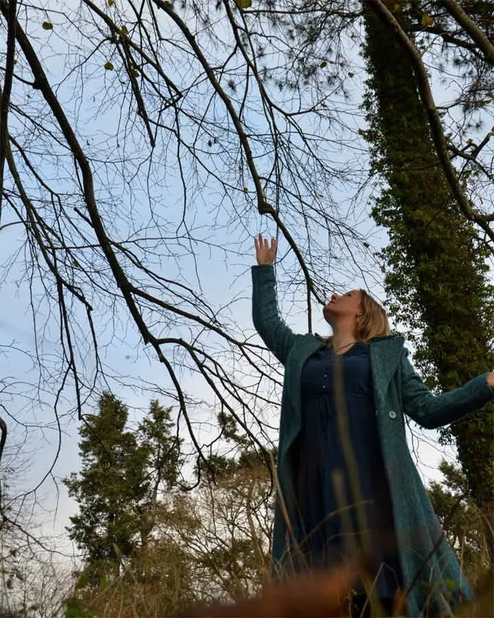 A woman standing outdoors reaching up towards a tree with leafless branches, wearing a teal coat and black dress, with trees and blue sky in the background.