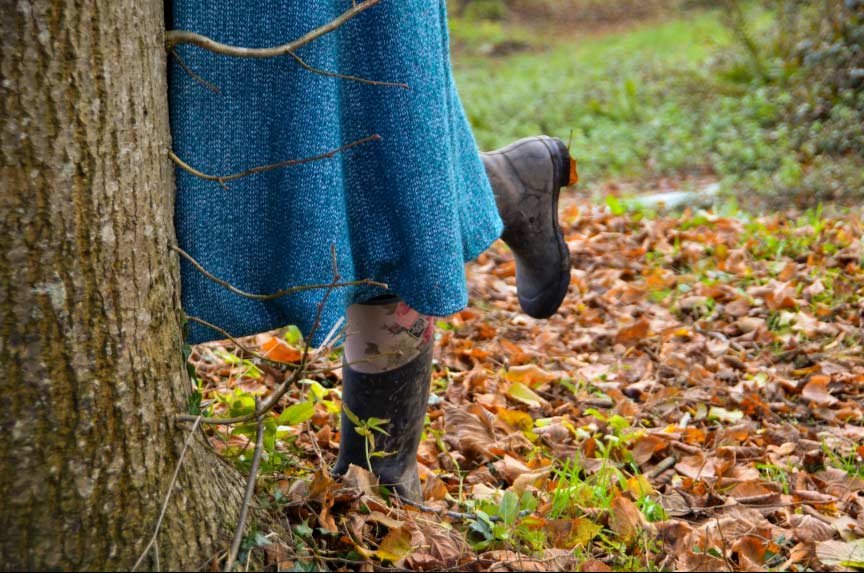 Close-up of a child's legs and feet, wearing rubber boots and blue tights, standing behind a tree in a park or forest with fallen autumn leaves and green grass on the ground.