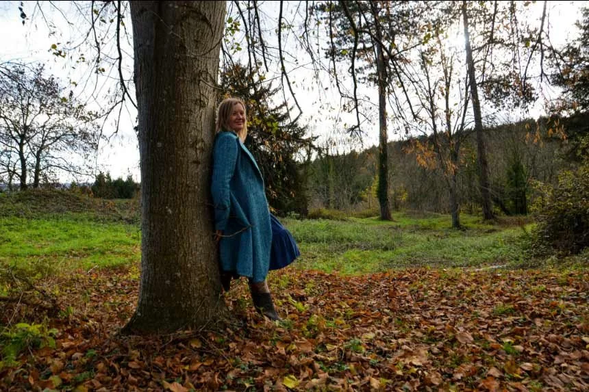 Woman in a blue coat standing against a large tree in a park during fall, with fallen leaves on the ground and trees with autumn foliage in the background.