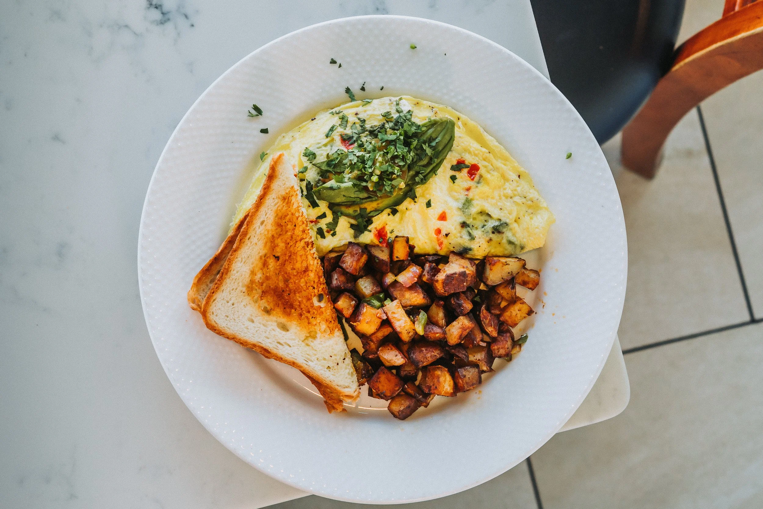 A white plate with scrambled eggs garnished with parsley, a serving of crispy hash browns, toasted bread, and garnished avocado slices.