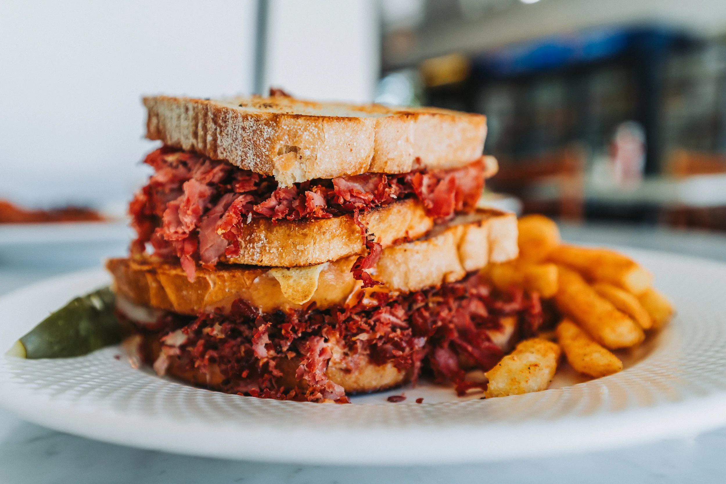 A plate with a sandwich made of sliced bread and piled up with pulled pork and shredded beef, with a side of French fries and a pickle