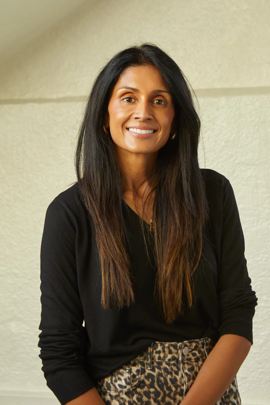 A woman with long dark hair smiling, wearing a black top and leopard print pants, standing against a light-colored textured wall.