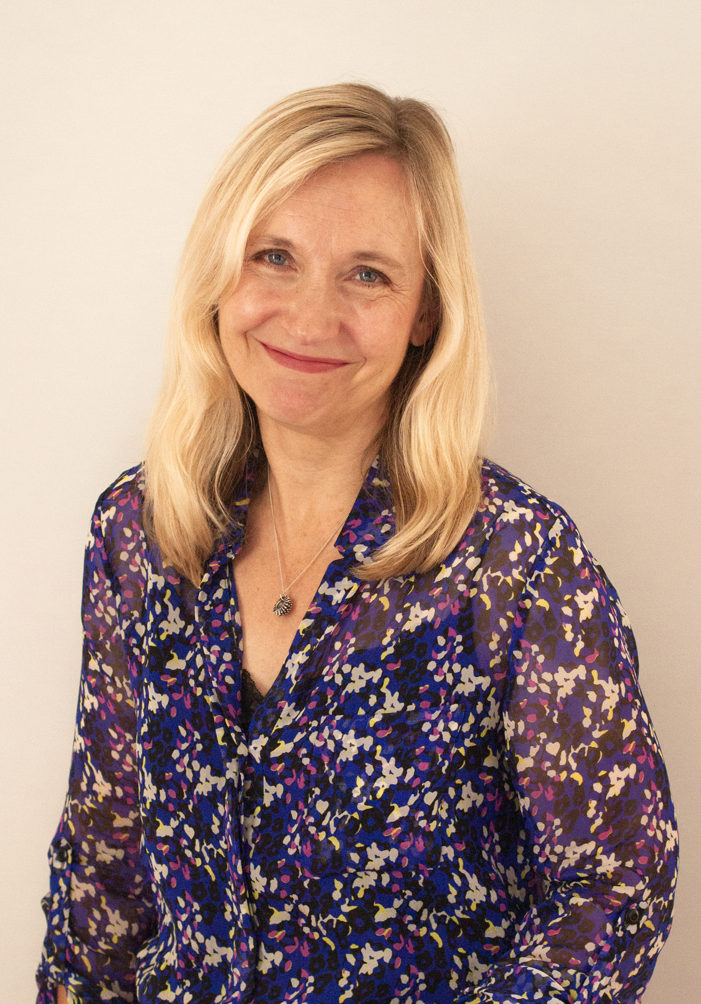 A woman with blonde hair smiling, wearing a colorful patterned blouse and a silver necklace, standing against a plain light-colored wall.