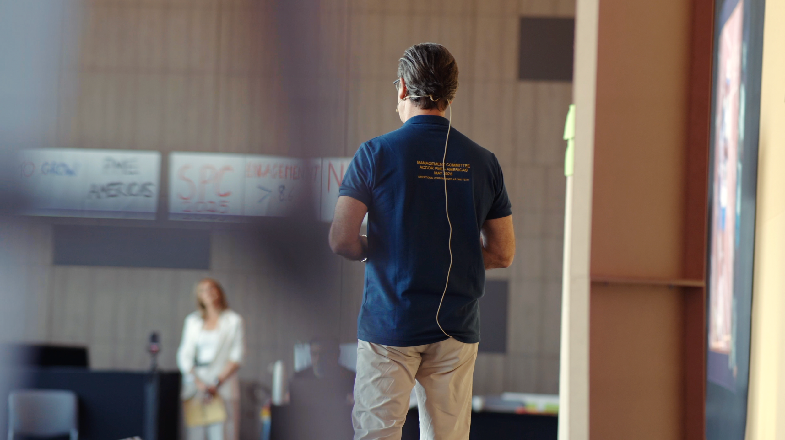 A woman is speaking at a conference about energy, with a presentation slide behind her that says 'Let's Talk About ENERGY.' Audience members are seated at tables, listening attentively.