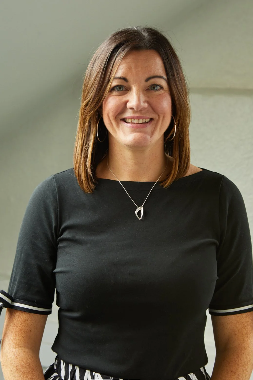 A woman with shoulder-length brown hair wearing a black top with striped sleeve cuffs, silver hoop earrings, and a silver necklace, smiling at the camera.