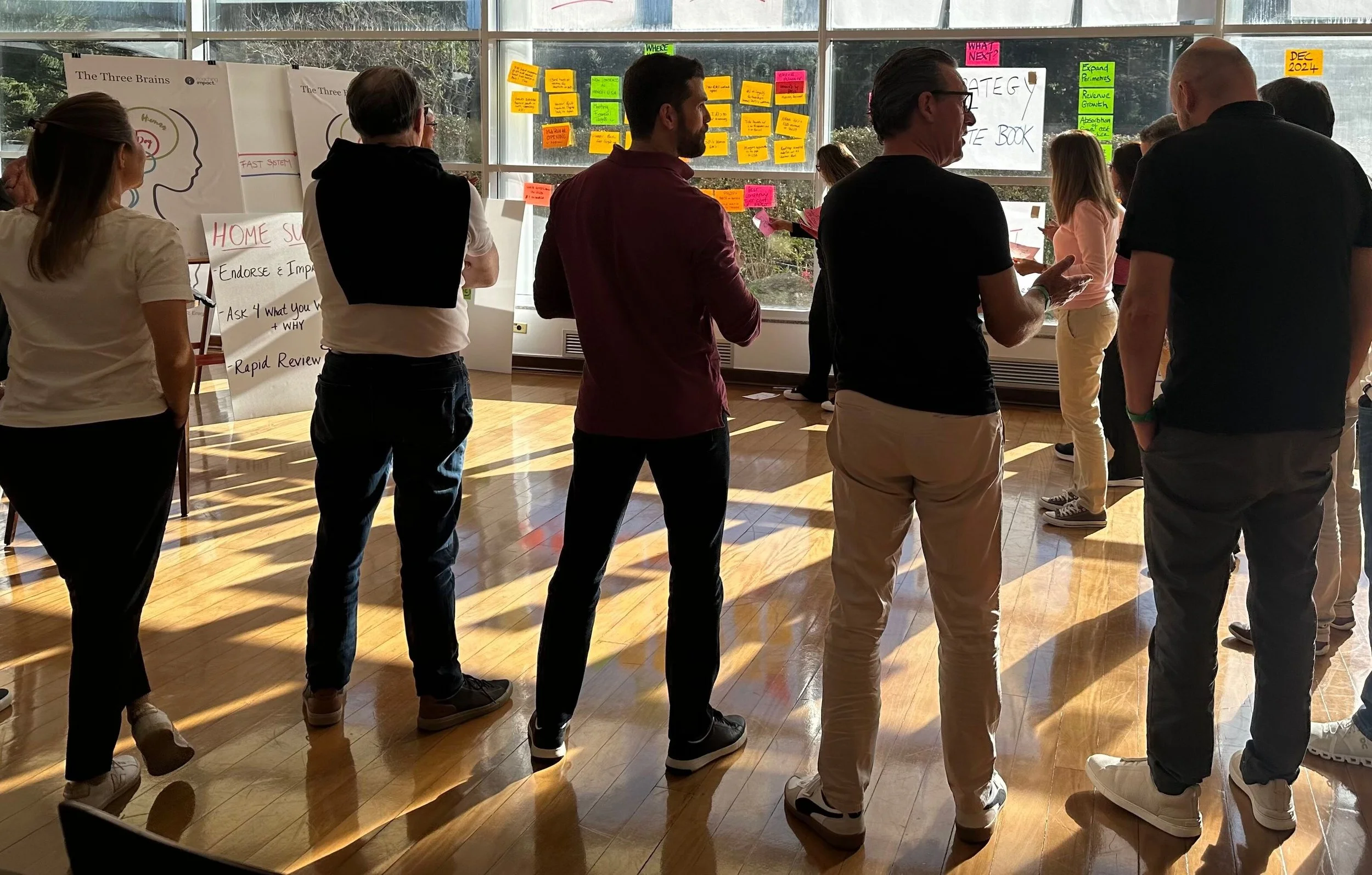 A group of people participating in a meeting or discussion in a modern office space. There are five individuals visible, with one person talking and others listening attentively. The setting includes a patterned black-and-white rug, coffee cups, notebooks, and a whiteboard on the wall.