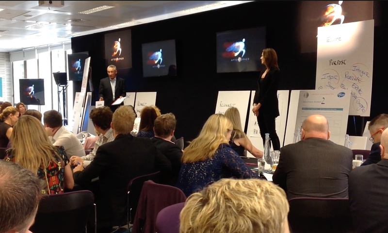 A group of people attending a business presentation or conference in a restaurant or hotel event space, with a speaker standing in front of a screen displaying a battery icon and text that says 'Energy Check'.