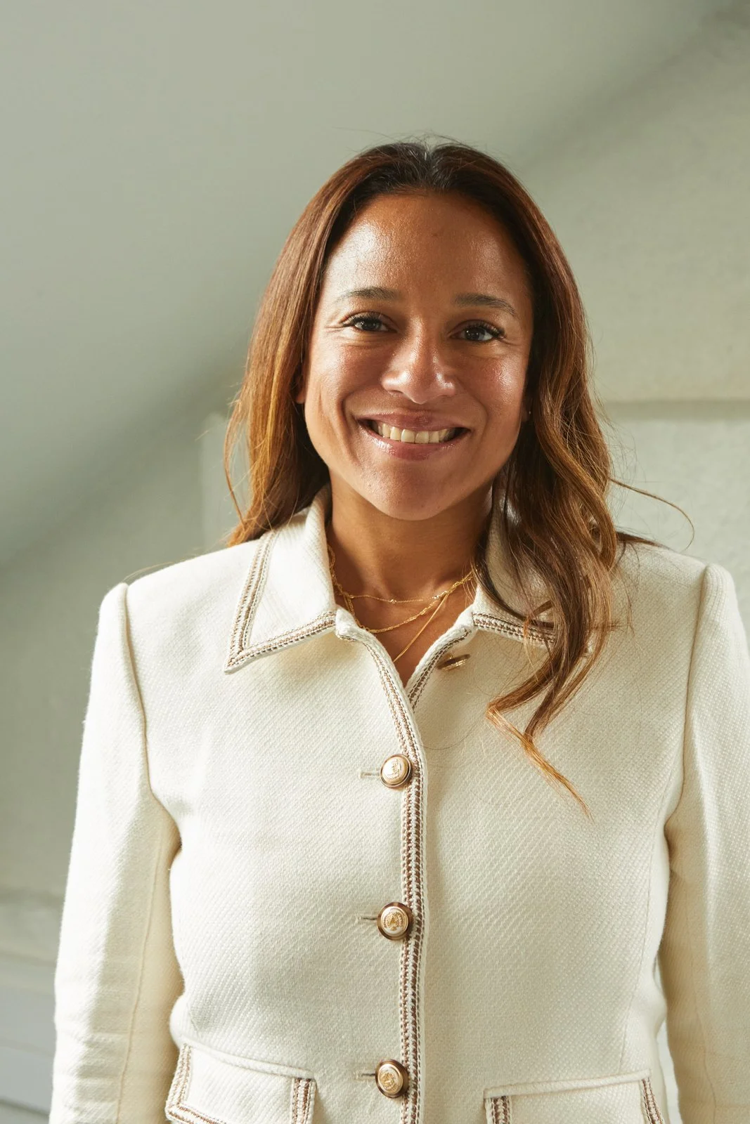 A woman with wavy brown hair smiling, wearing a white jacket with gold buttons and a layered gold necklace, standing indoors against a neutral background.