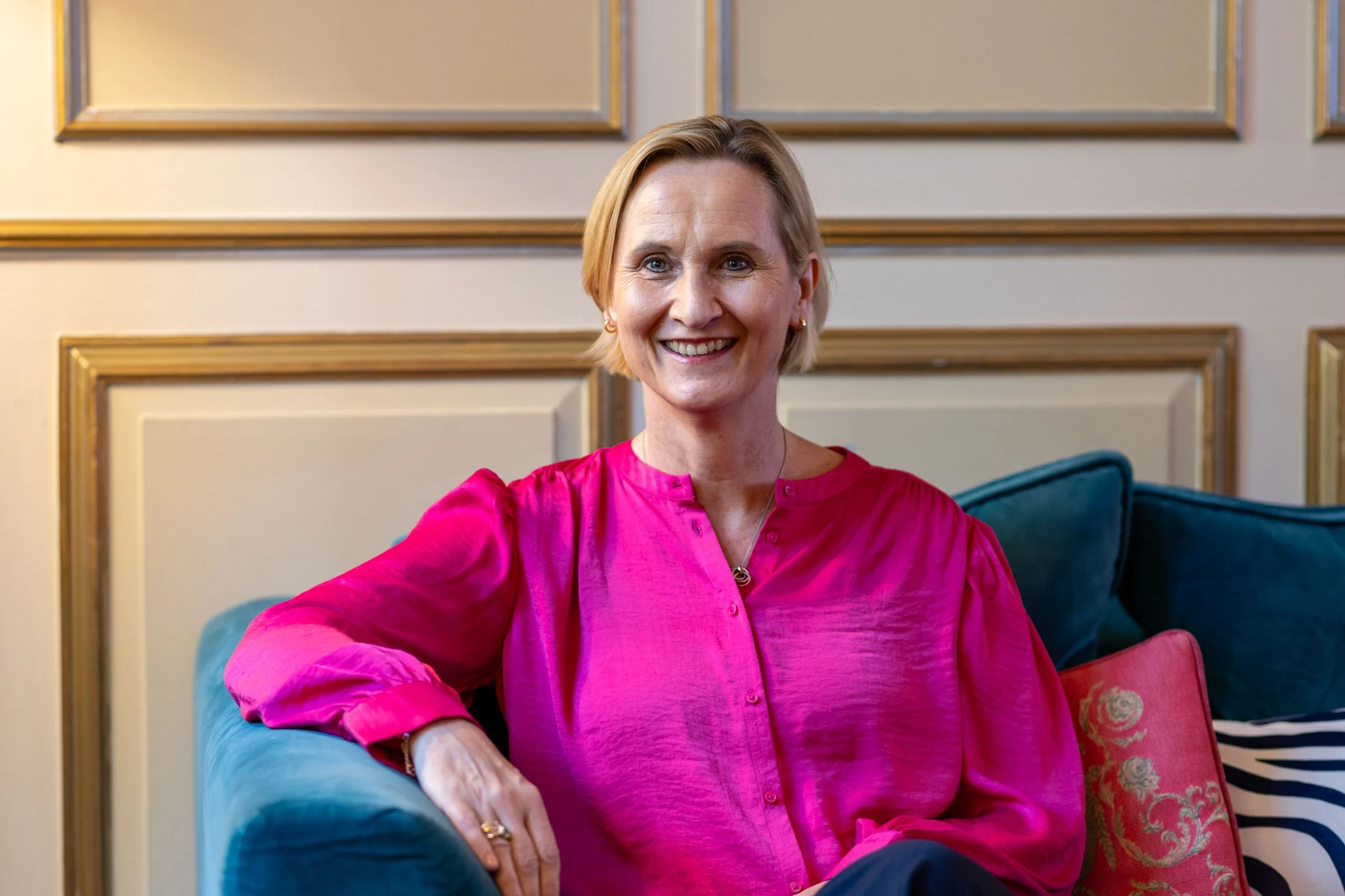 A woman with short blonde hair wearing a bright pink blouse sitting on a teal couch in a room with cream-colored walls and gold trim, smiling at the camera.