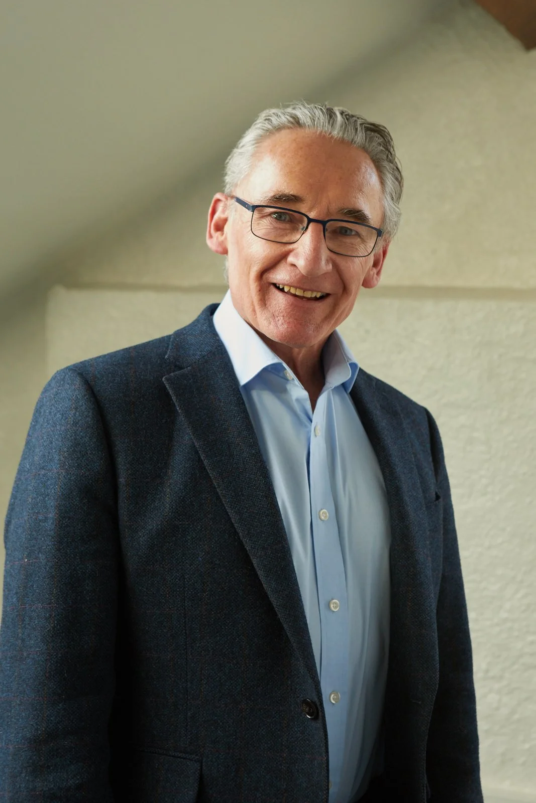 Older man with gray hair, glasses, and a suit smiling indoors.