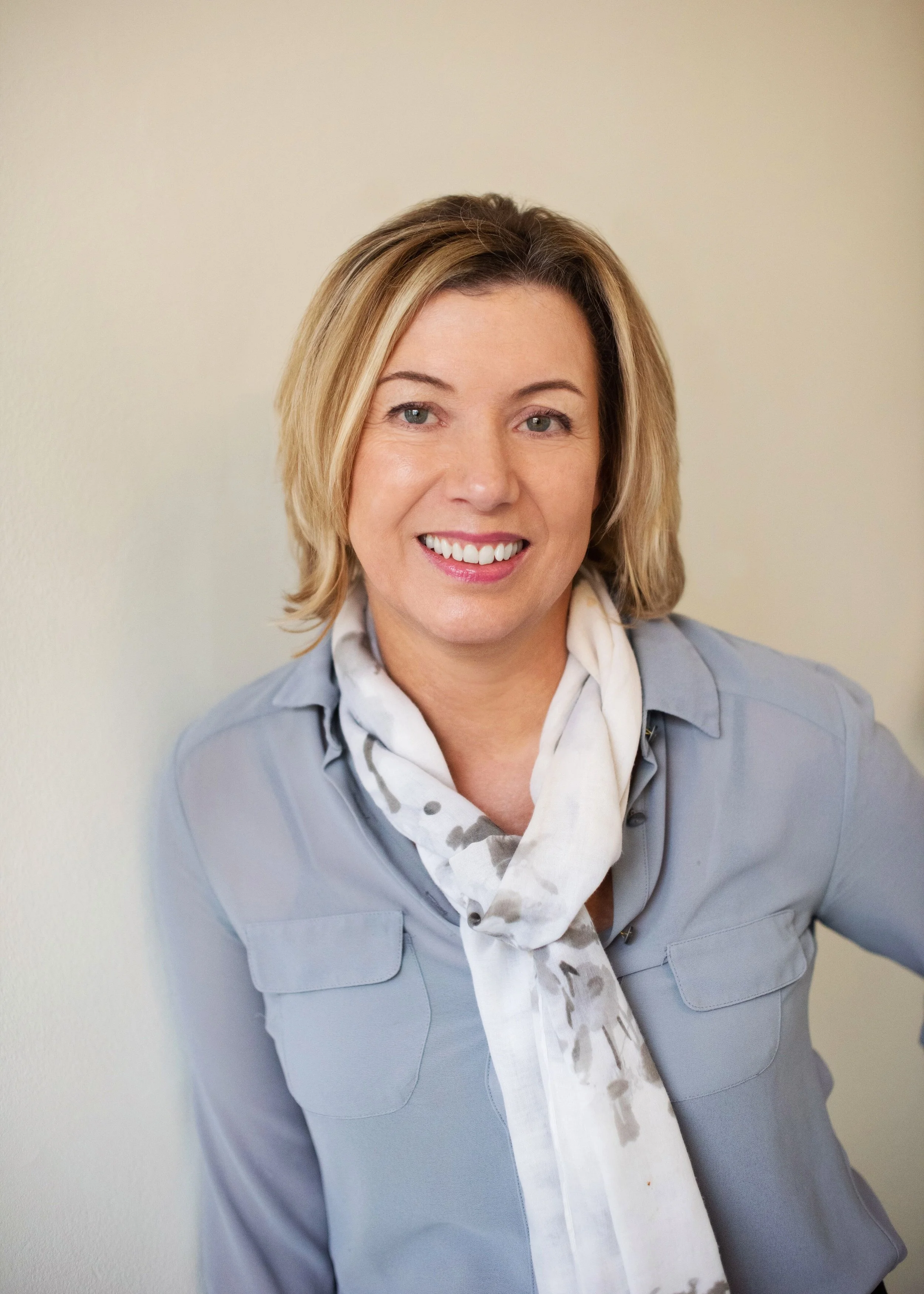 Smiling woman with shoulder-length blonde hair wearing a beige sweater standing indoors with a light-colored wall in the background.
