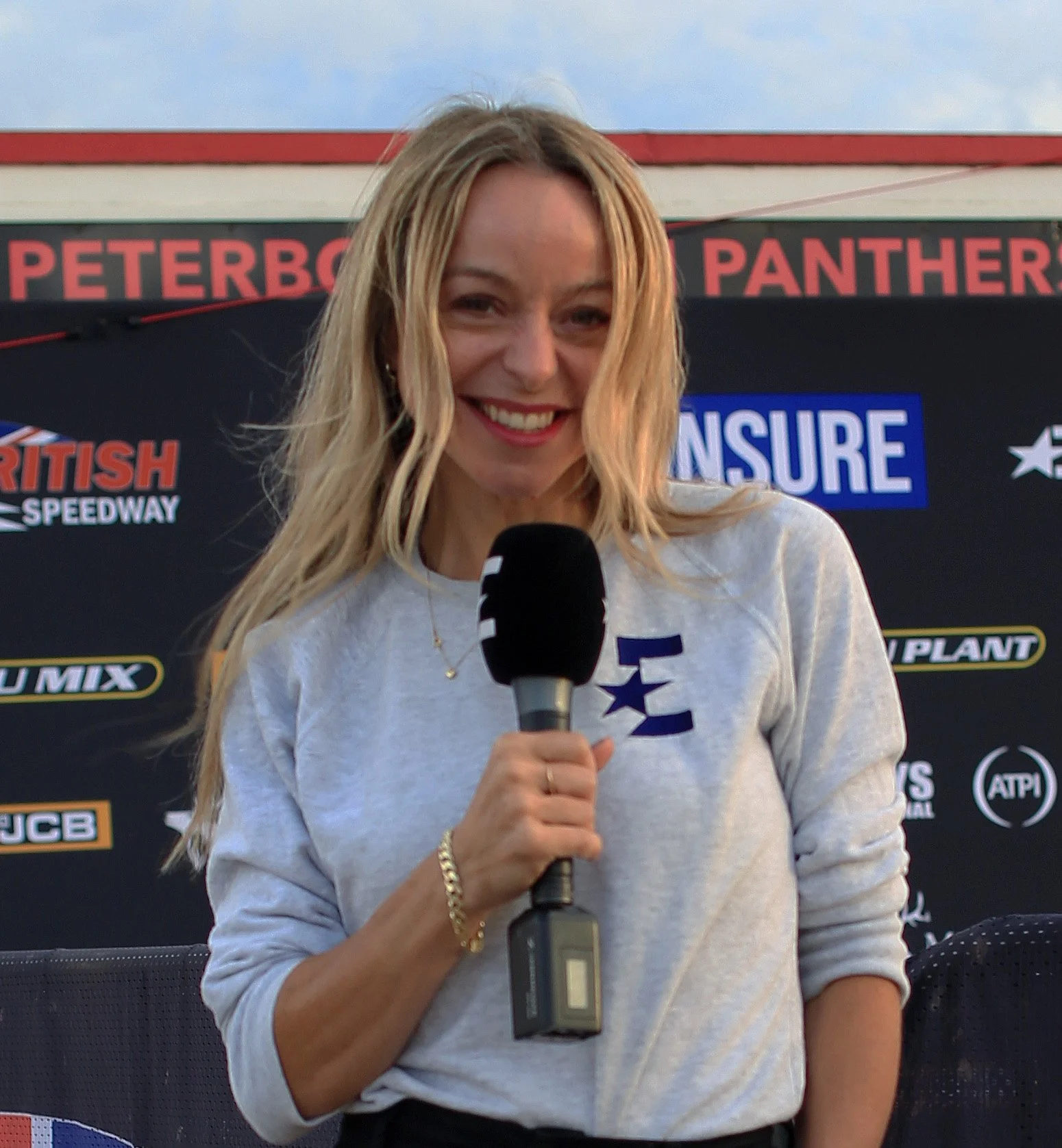 A woman with blonde hair holding a microphone, smiling, standing in front of a black backdrop with logos and text, at what appears to be a racing event at British Speedway.