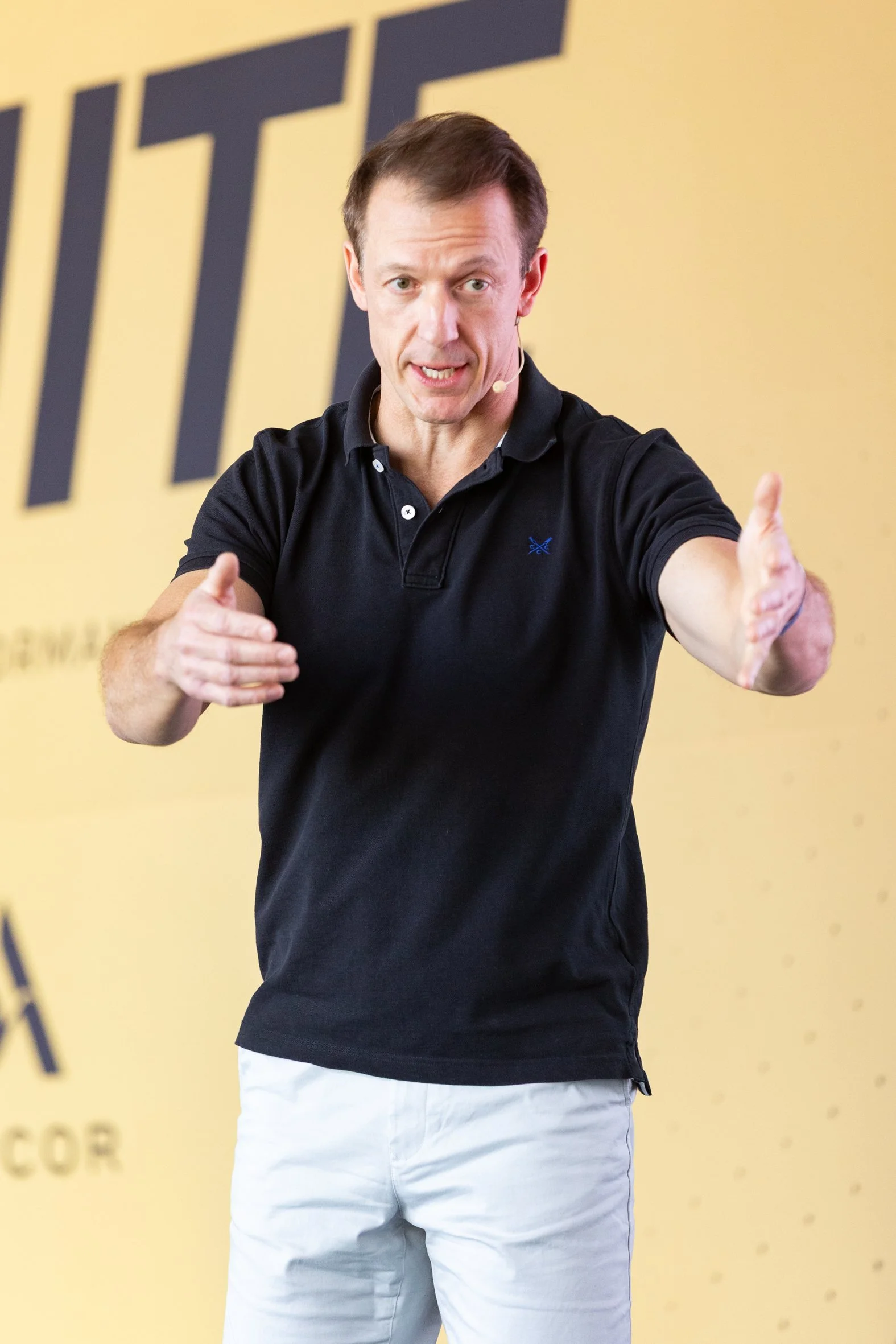 A man in a black polo shirt and white pants speaking on stage with a yellow background and black lettering.