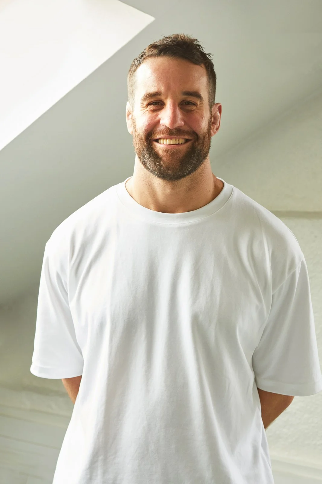 Smiling man with beard and short hair wearing a white t-shirt, standing indoors.
