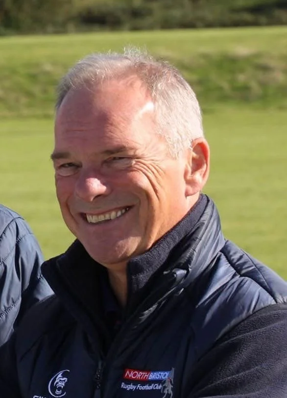 A middle-aged man with short light-colored hair, smiling, wearing a dark blue knitted sweater over a collared shirt, standing against a neutral light-colored wall.