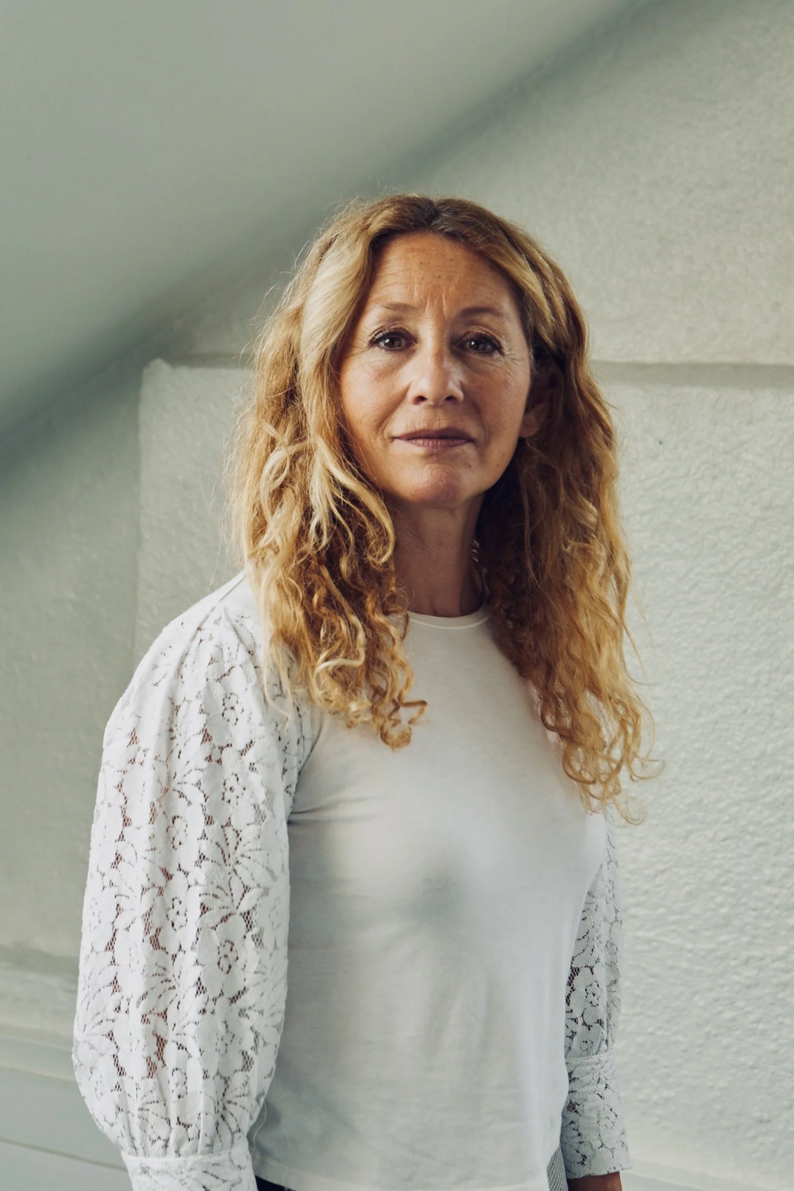 A woman with curly red hair wearing a white top with lace sleeves, standing in a room with a textured light green wall.